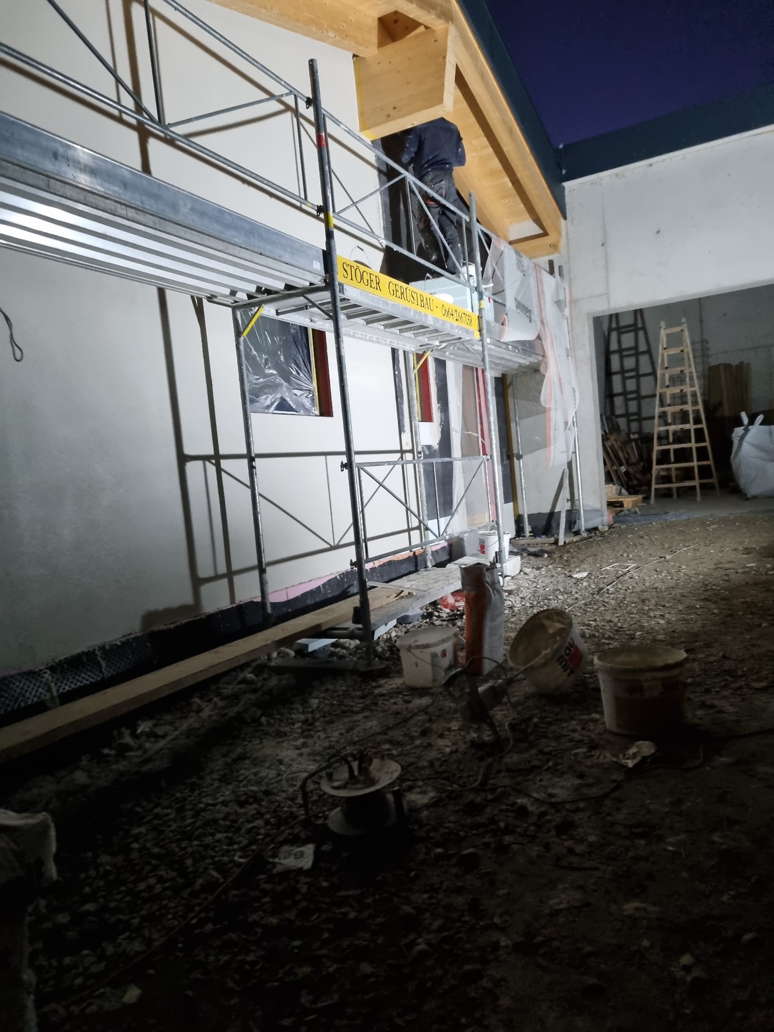 Elewacja budynku w trakcie prac, widoczne rusztowanie z pracownikiem, okno zabezpieczone folią, materiały budowlane na ziemi, drewniana konstrukcja dachu.