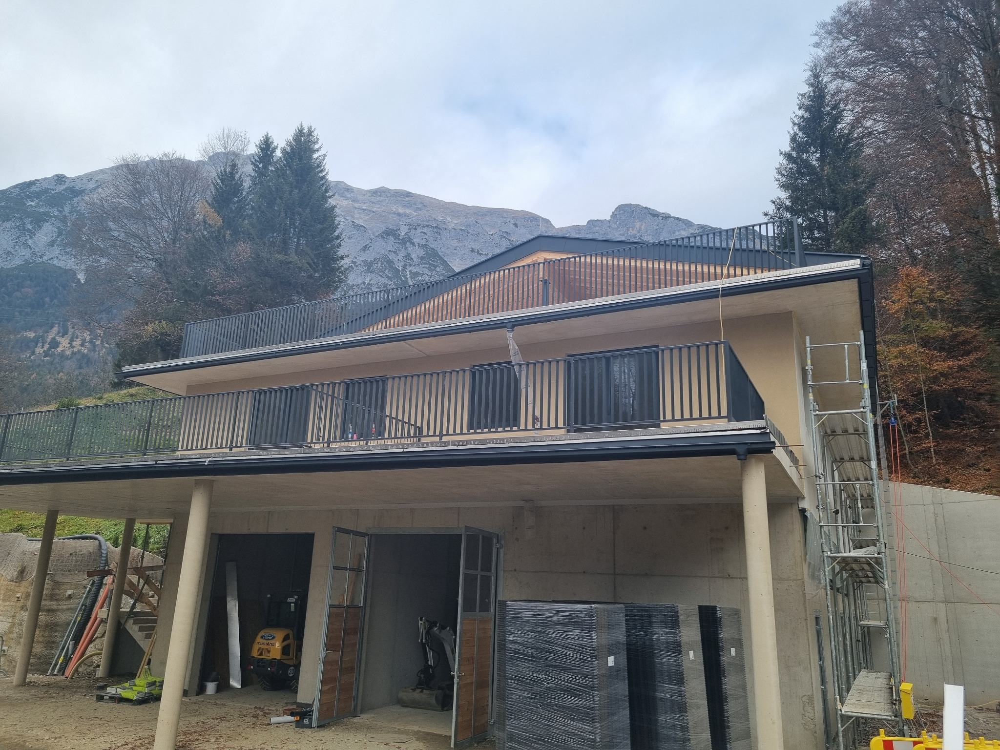 Dwukondygnacyjny dom w trakcie budowy z widoczną elewacją, balkonami z metalowymi balustradami i częściowo otwartym parterem, na tle górskiego krajobrazu.