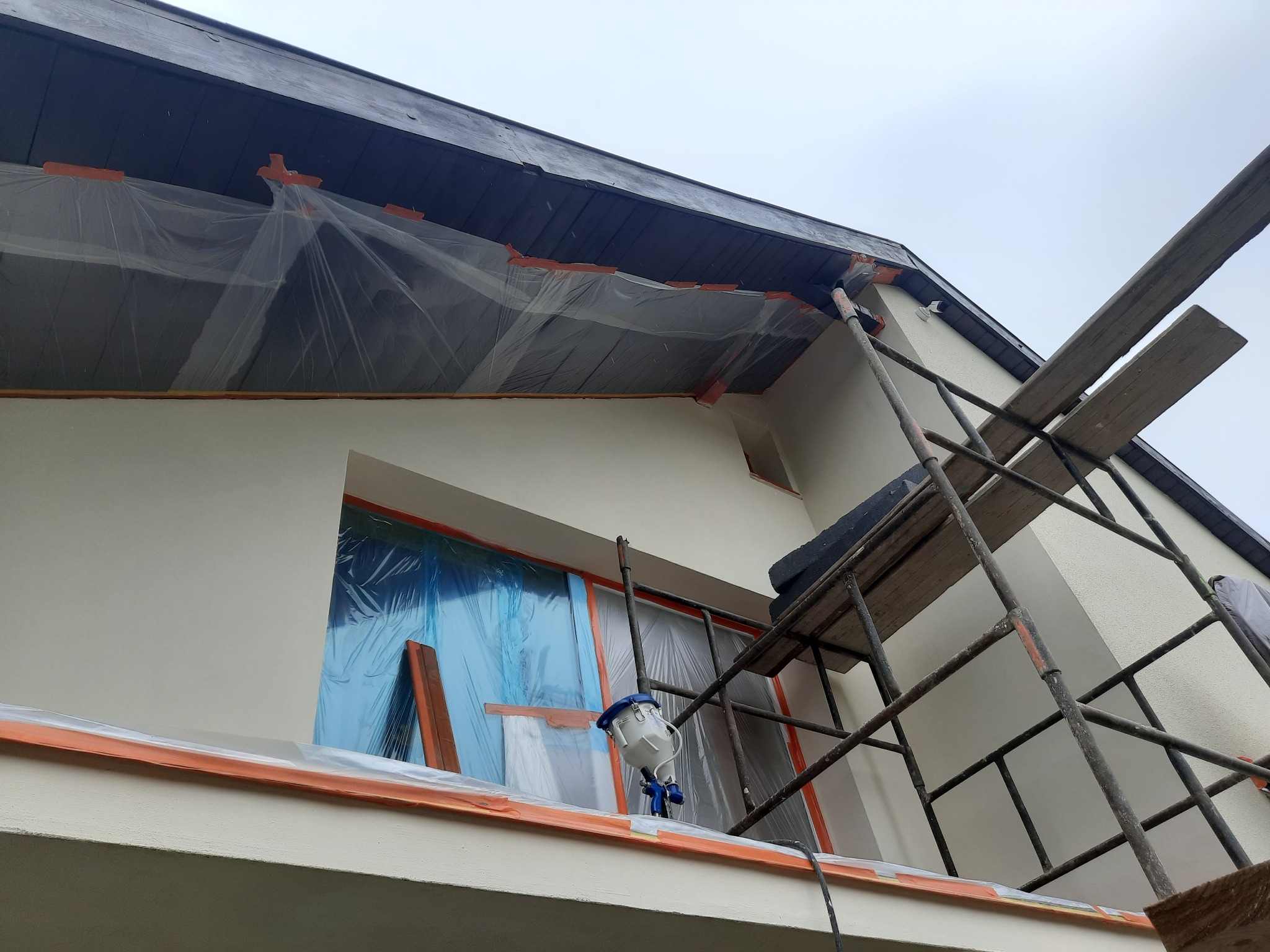 Elewacja domu zabezpieczona folią i taśmą malarską przed natryskowym malowaniem, widoczny pistolet natryskowy, rusztowanie i deski.