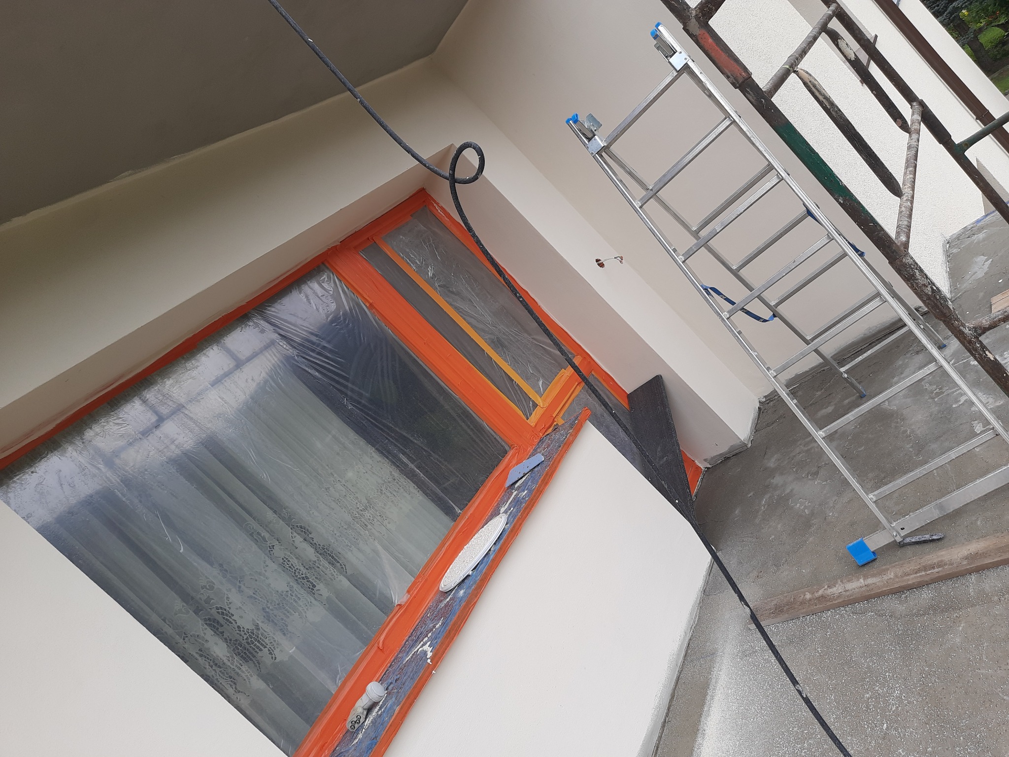 Ocieplona elewacja budynku w trakcie malowania, okno zabezpieczone folią, widoczna drabina i kabel od agregatu malarskiego.
