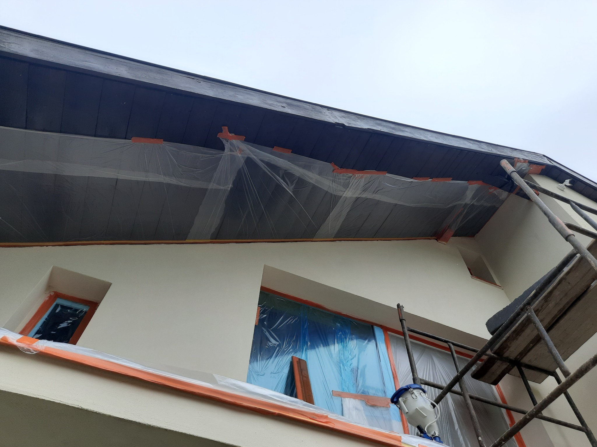 Elewacja domu zabezpieczona folią i taśmą malarską przed malowaniem, widoczne rusztowanie i urządzenie do malowania natryskowego.