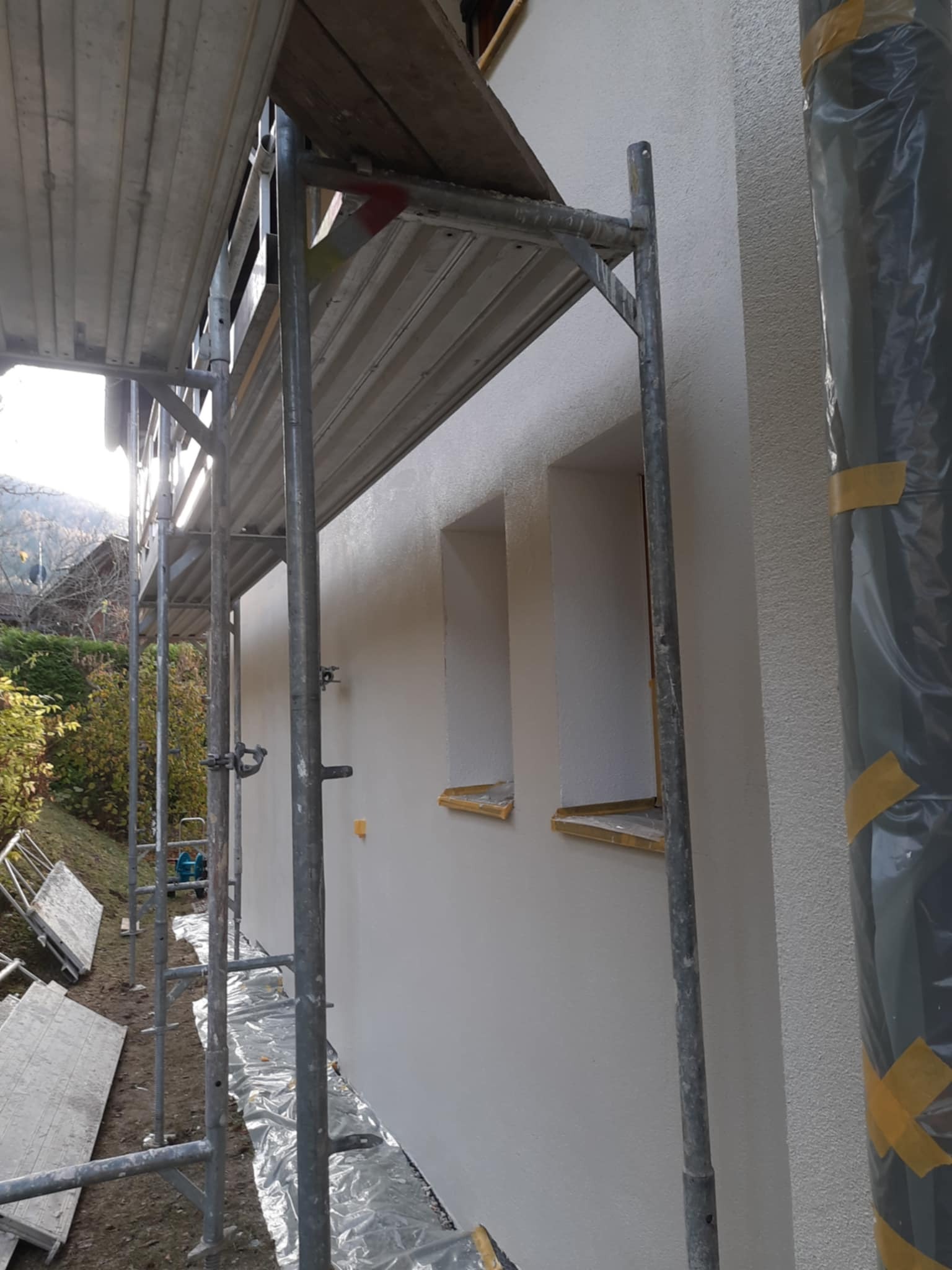 Świeżo otynkowana elewacja domu jednorodzinnego, widok z rusztowania, okna zabezpieczone folią i taśmą malarską, w tle zieleń ogrodu.