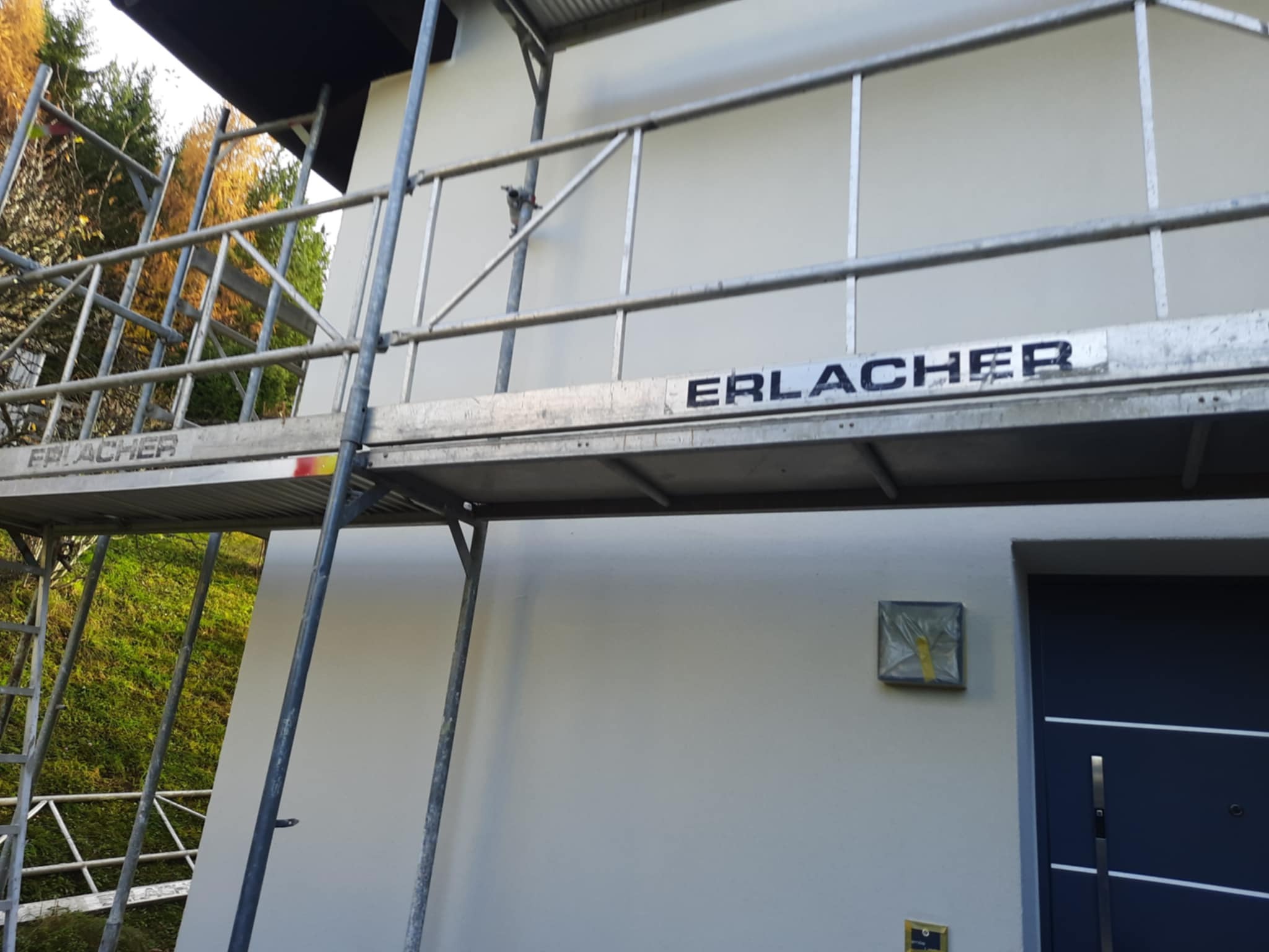 Świeżo otynkowana elewacja domu jednorodzinnego z szarymi detalami, widoczna z poziomu rusztowania firmy Erlacher, z zabezpieczonym oknem i fragmentem ciemnych drzwi.