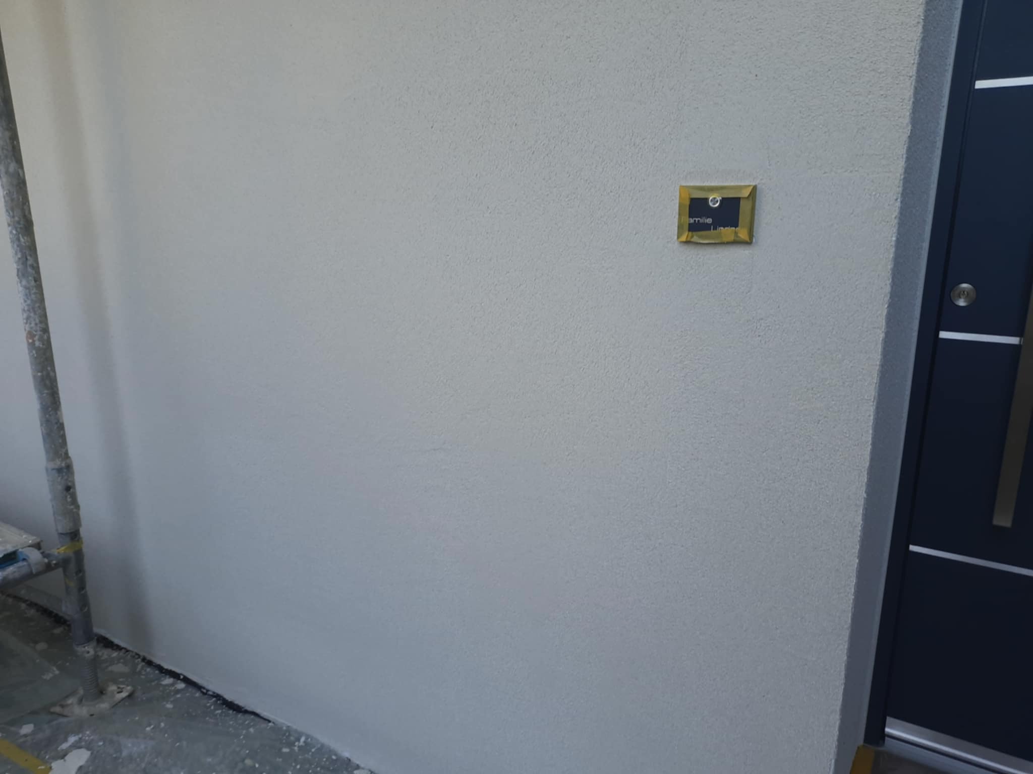 Świeżo otynkowana, jasnoszara elewacja z widoczną fakturą, fragment rusztowania po lewej i ciemne drzwi wejściowe po prawej, mała tabliczka informacyjna na ścianie.