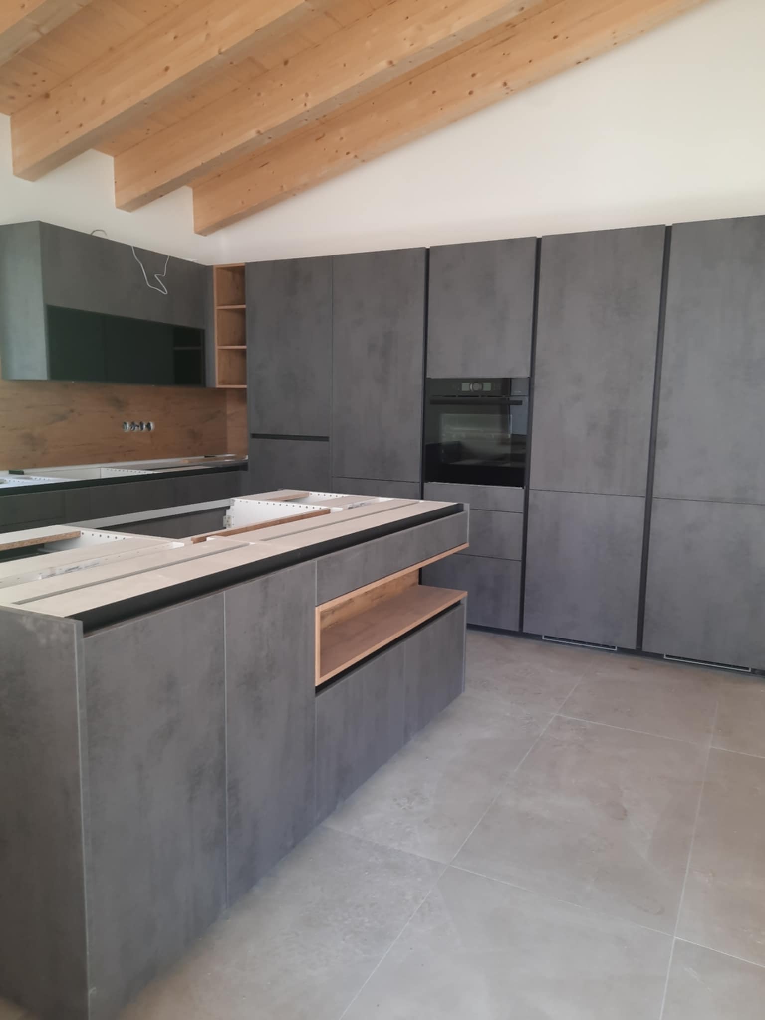 Wnętrze kuchni w trakcie montażu: szare, minimalistyczne szafki kuchenne z drewnianymi akcentami, wyspa kuchenna w stanie surowym, widoczne drewniane belki stropowe.