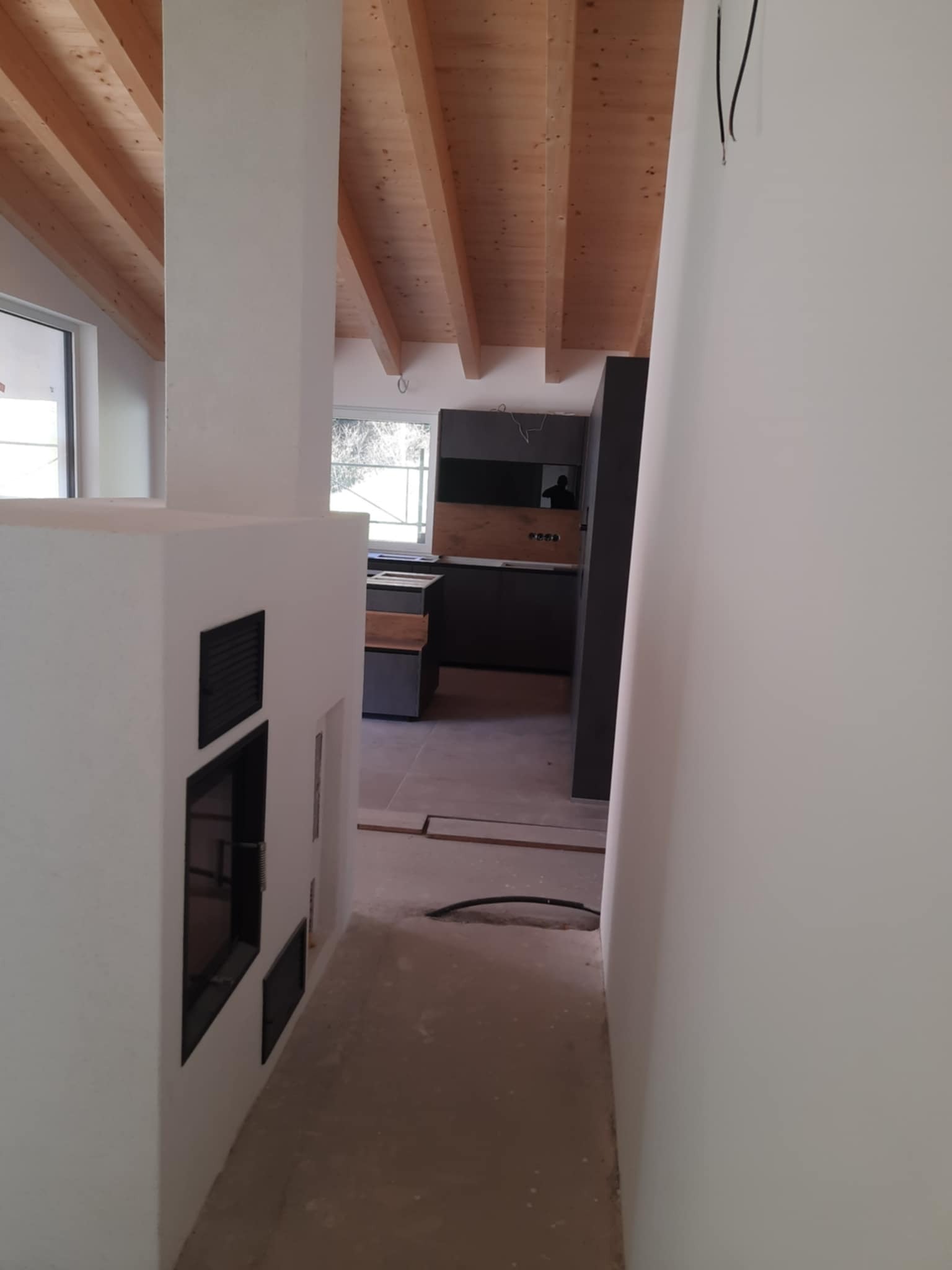 Wnętrze domu w trakcie wykańczania, widoczna surowa podłoga, ściany przygotowane do malowania, drewniany strop oraz fragment zabudowy kominkowej i kuchni z ciemnymi szafkami.