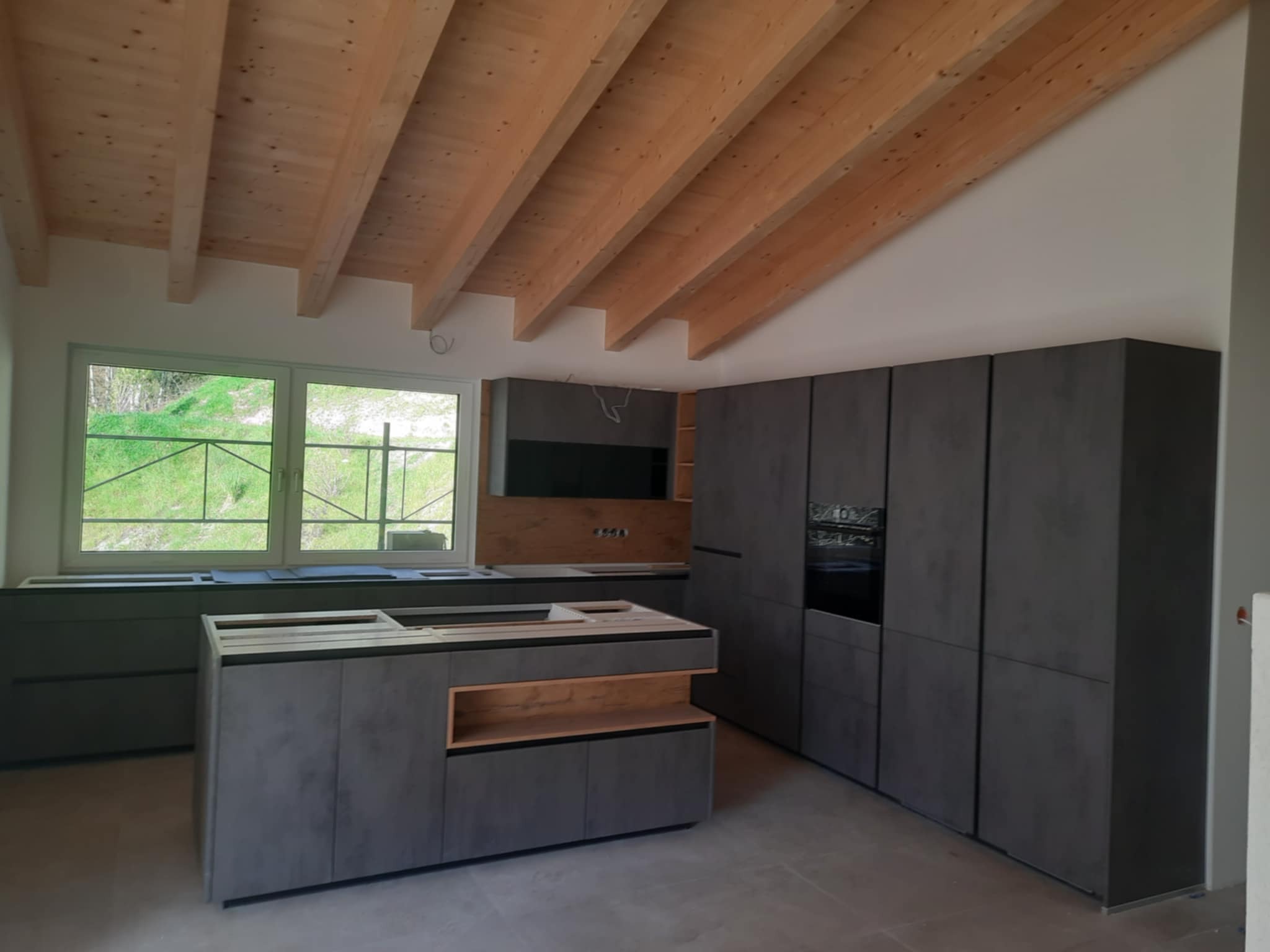 Wnętrze kuchni w trakcie montażu: szare, matowe szafki kuchenne z drewnianymi akcentami, wyspa kuchenna bez blatu, widoczne okno z zielenią na zewnątrz, surowy drewniany strop z belek.
