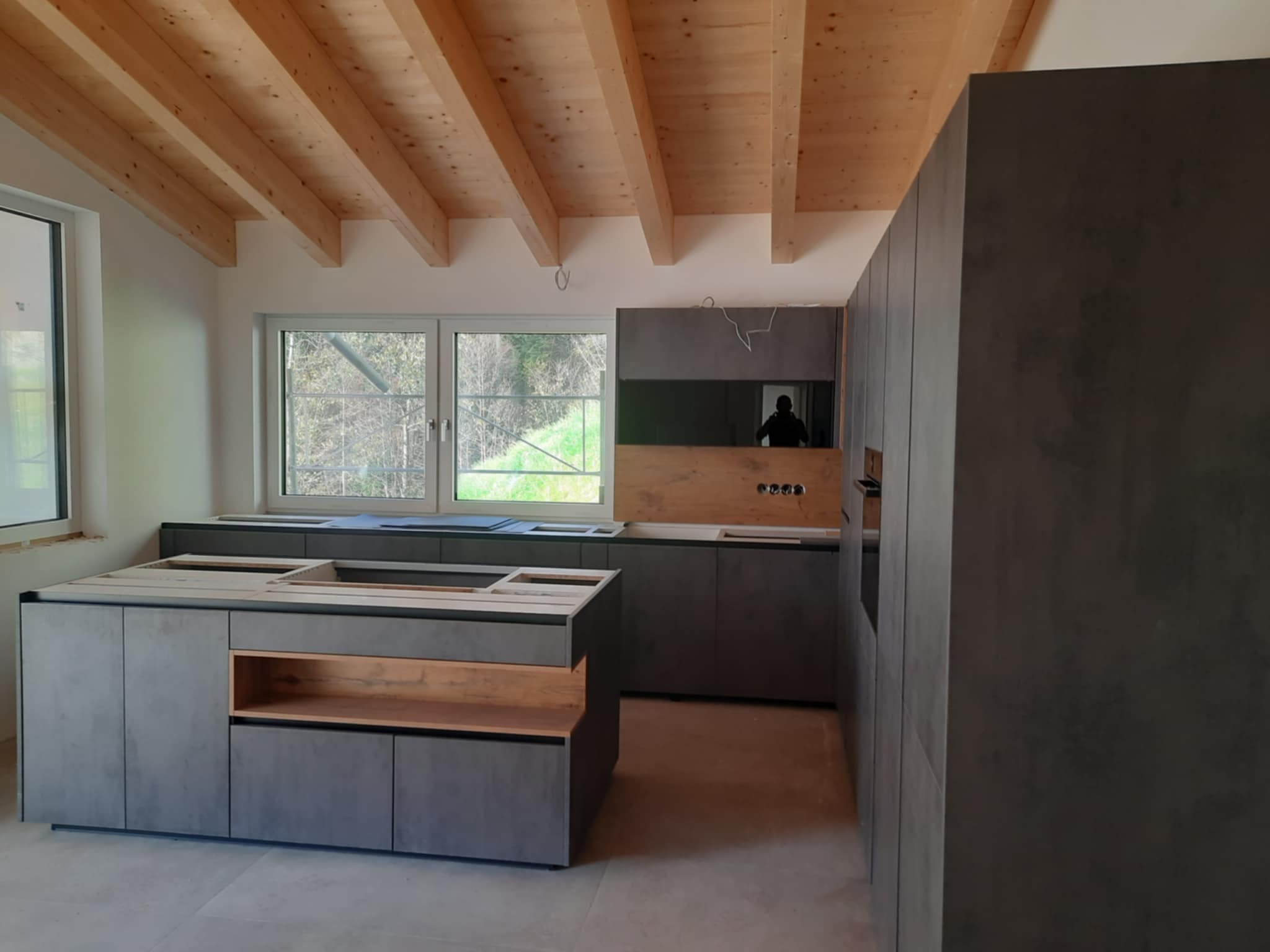 Surowa kuchnia z szarymi szafkami i drewnianymi akcentami, widoczne surowe drewniane belki stropowe i okno z widokiem na zieleń, w trakcie montażu, bez blatu.