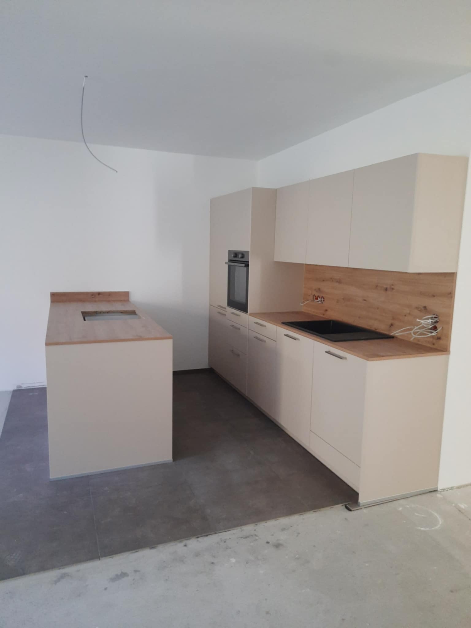 Niedokończona kuchnia w jasnych barwach z drewnianym blatem i wyspą, widoczne wystające kable i surowy beton na podłodze.