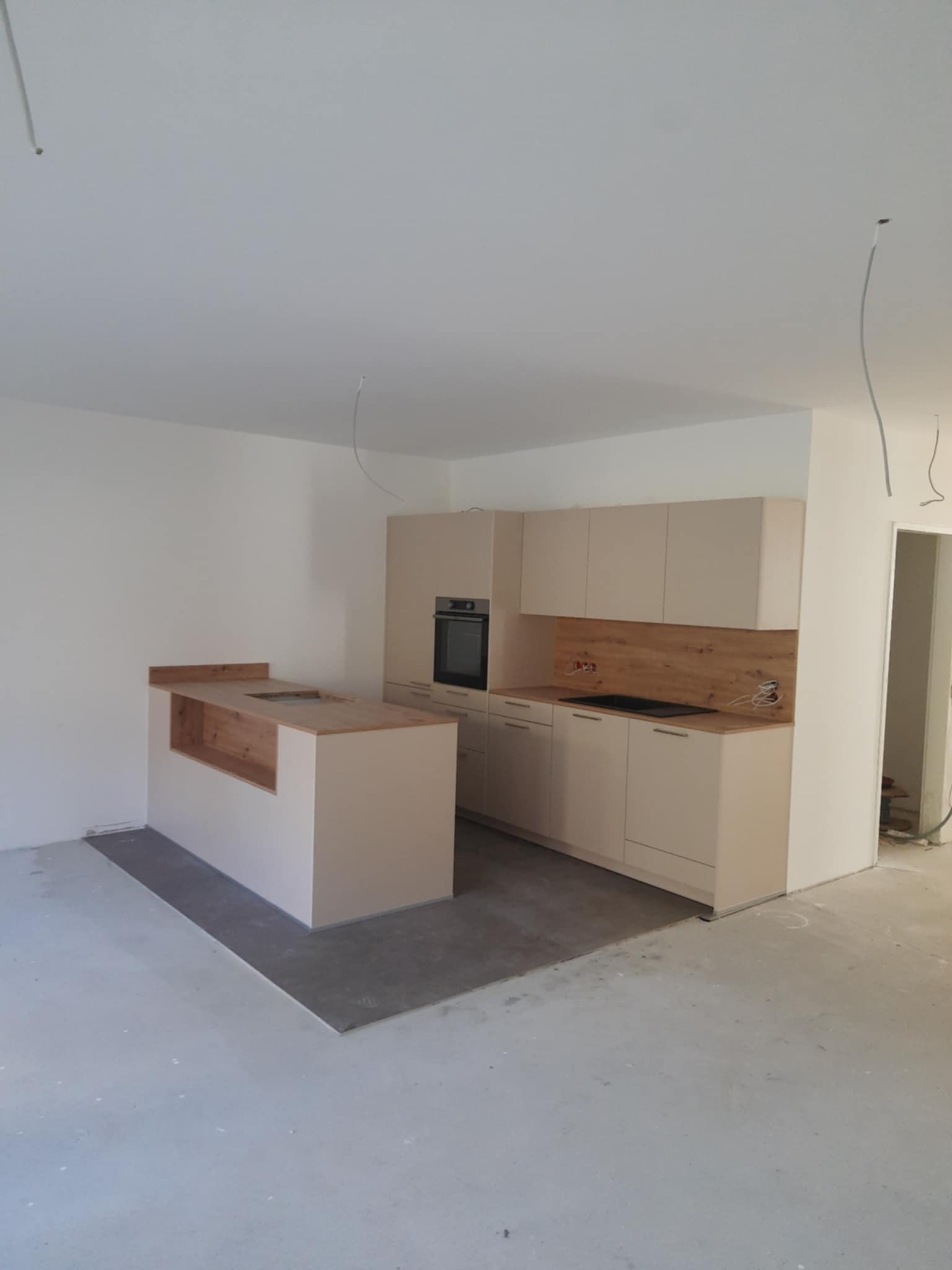 Wnętrze kuchni w trakcie wykańczania w Kłodzku: beżowe szafki kuchenne z drewnianym blatem, wyspa kuchenna z surowym wykończeniem, widoczne kable zwisające z sufitu, szara podłoga wylewka.