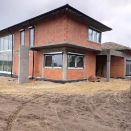 Surowy stan budynku dwukondygnacyjnego z czerwonej cegły z widocznymi elementami żelbetowymi, osadzony na piaszczystym terenie z wyraźnymi śladami opon.