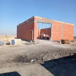 Wznoszenie ścian budynku z czerwonej cegły, widoczne stalowe słupy podtrzymujące nadproże otworu, palety z cegłami i sprzęt budowlany w tle.