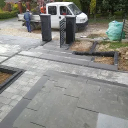 Schody i ścieżka z szarej kostki brukowej prowadzące do furtki z czarnymi słupkami, w tle biały samochód dostawczy i pracownik w niebieskiej koszuli.