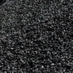 Góra czarnego ekogroszku z widocznymi oponami pojazdu w tle, lekko oświetlona.