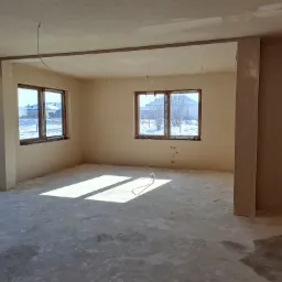 Wnętrze surowego domu po nałożeniu tynków, widoczne gładkie ściany w odcieniach beżu, surowy beton na podłodze oraz wystające przewody elektryczne z sufitu, w tle okna z widokiem na zaśnieżone...