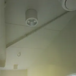 Cylindryczna lampa sufitowa w nowo wykończonym wnętrzu, widoczne rury instalacyjne i fragment wentylacji, naturalne światło wpadające przez okno.