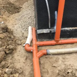 Pomarańczowe rury kanalizacyjne ułożone w wykopie, częściowo owinięte geowłókniną, widoczna ściana fundamentowa z izolacją kubełkową i koparka w tle.