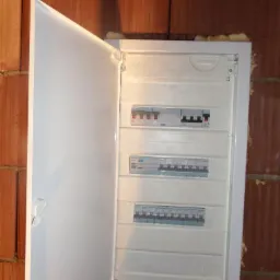 Otwarta skrzynka elektryczna z widocznymi bezpiecznikami zamontowana na ścianie z czerwonej cegły. Widoczne ślady pianki montażowej wokół obudowy.