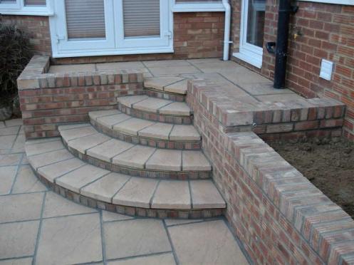 Zewnętrzne schody z cegły klinkierowej i jasnych płytek, prowadzące do drzwi wejściowych, z widoczną instalacją odprowadzającą wodę deszczową.