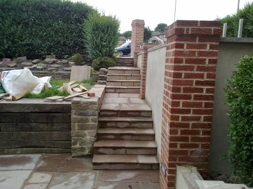 Kamienne schody ogrodowe z ceglanymi słupkami, widok z dołu, częściowo ukończone, z workiem i drewnem obok, widoczne ślady wilgoci na kamieniach