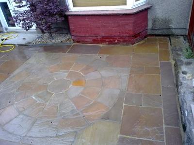 Ułożony wzór z płytek chodnikowych w kształcie okręgu, z centralnym żółtym elementem, przylegający do ceglanej elewacji budynku, obok rośnie purpurowy krzew.