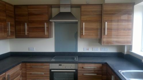 Nowoczesna kuchnia z drewnianymi szafkami z widocznym usłojeniem, czarnym blatem i srebrnymi uchwytami, okap kuchenny nad płytą grzewczą, białe ściany z gniazdkami elektrycznymi.