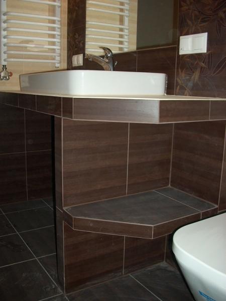 Łazienka z brązowymi płytkami imitującymi drewno, biała umywalka nablatowa i grzejnik drabinkowy, widoczna półka z płytek pod umywalką.