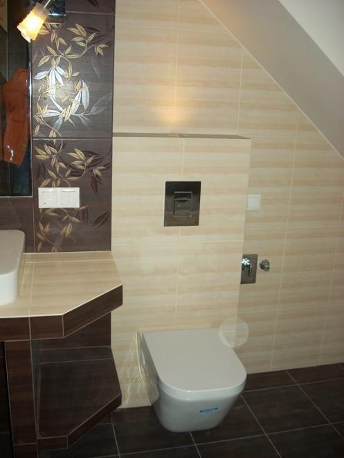 Jasna łazienka na poddaszu z beżowymi płytkami imitującymi kamień, dekoracyjnymi brązowymi płytkami z motywem roślinnym, podwieszaną toaletą i ciemnobrązową podłogą z płytek.