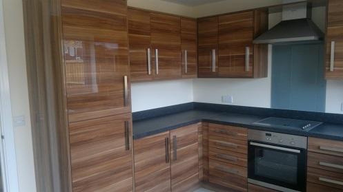 Nowoczesna kuchnia z frontami szafek w odcieniach orzecha włoskiego, czarnym blatem i srebrnymi uchwytami, widok na piekarnik i okap.