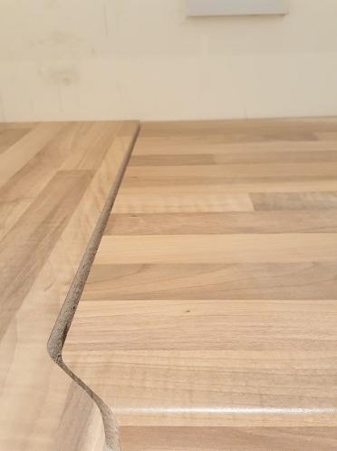 Fragment dwóch połączonych, jasnych blatów drewnianych, jeden z zaokrągloną krawędzią, widoczne delikatne uszczelnienie między nimi, na neutralnym tle.