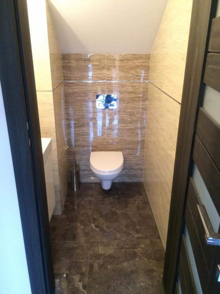 Wąska toaleta z brązowymi płytkami imitującymi drewno na ścianach i podłodze, podwieszana muszla klozetowa z chromowanym przyciskiem spłukującym, widok zza otwartych, ciemnych drzwi.