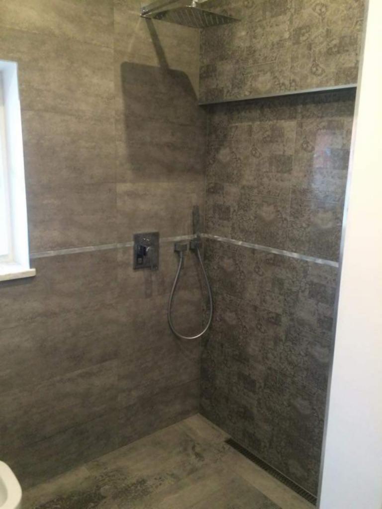 Wykończona kabina prysznicowa z szarymi płytkami o wzorzystej fakturze, deszczownicą i odpływem liniowym w podłodze, widok z narożnika.