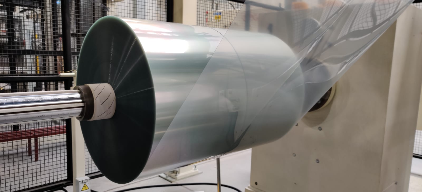 Duża rolka przezroczystej folii nawinięta na metalowy wałek z gumowymi pierścieniami na maszynie przemysłowej w hali produkcyjnej.