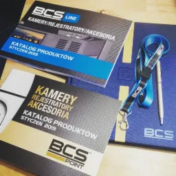 Katalogi produktowe BCS Line i BCS Point z kamerami, rejestratorami i akcesoriami, leżące obok notatnika i smyczy z logo BCS, całość w tonacji niebiesko-szaro-czarnej.