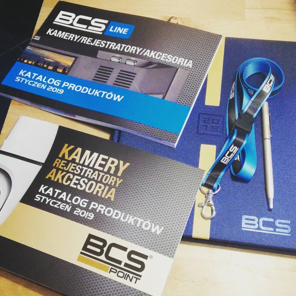 Katalogi produktowe BCS Line i BCS Point z kamerami, rejestratorami i akcesoriami, leżące obok notatnika i smyczy z logo BCS, całość w tonacji niebiesko-szaro-czarnej.
