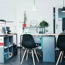 Jasne biuro z biurkami, krzesłami i komputerem. Na ścianie widoczny zegar i półki z roślinami. Minimalistyczny design.