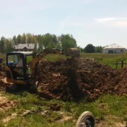 Żółta mini koparka w trakcie prac ziemnych w Białymstoku, z widoczną stertą ziemi i pracownikami w tle, krajobraz z domami w oddali.