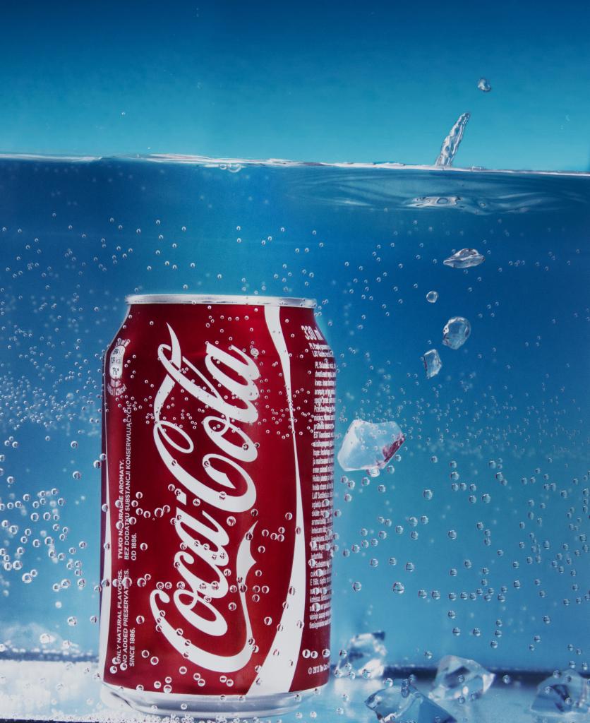 Puszka Coca-Coli zanurzona w wodzie z kostkami lodu, bąbelki powietrza otaczają puszkę, a nad nią widać powierzchnię wody i kroplę.
