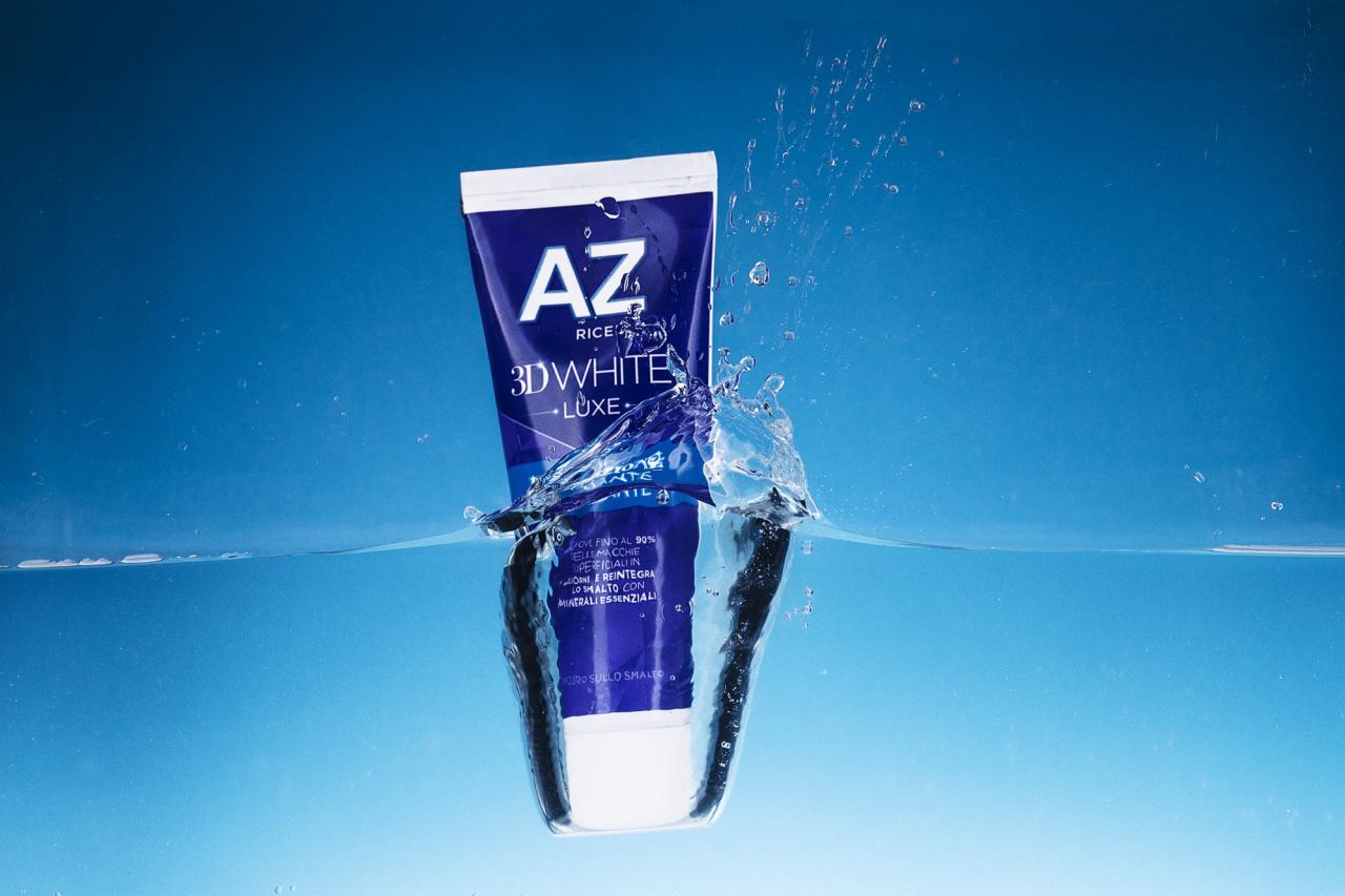 Niebieska tuba pasty do zębów AZ 3D White Luxe częściowo zanurzona w czystej wodzie, z rozpryskującymi się kroplami na tle jasnoniebieskiego gradientu.