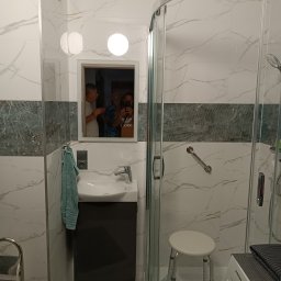 Usługi remontowo budowlane Jakub Justynski - Wykończona łazienka z kabiną prysznicową, umywalką z szafką w kolorze antracytowym, biało-szarymi płytkami imitującymi marmur i akcentem z płytek w odcieniach zieleni, z widocznym odbiciem trzech...