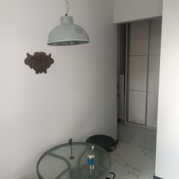 Usługi remontowo budowlane Jakub Justynski - Szklany stół z metalową ramą i dwoma czarnymi stołkami w rogu pokoju z marmurową podłogą, wisząca szara lampa nad stołem, ozdobny element na ścianie i biała szafa z przesuwnymi drzwiami w tle.