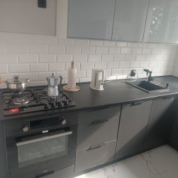 Usługi remontowo budowlane Jakub Justynski - Nowoczesna, szara kuchnia z białymi płytkami typu metro, czarnym blatem i sprzętem AGD: piekarnik Amica, płyta gazowa, czarny zlew i bateria. Na blacie czajnik i kawiarka.