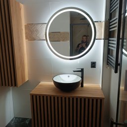 Usługi remontowo budowlane Jakub Justynski - Nowoczesna łazienka z okrągłym lustrem z podświetleniem LED, czarną umywalką nablatową i szafką z lamelami w kolorze drewna, widoczny grzejnik drabinkowy i fragment kabiny prysznicowej.
