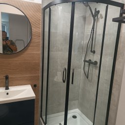 Usługi remontowo budowlane Jakub Justynski - Nowoczesna łazienka z narożną kabiną prysznicową z czarnymi profilami, umywalką z szafką w kolorze granatowym i okrągłym lustrem, odbijającym osobę robiącą zdjęcie.