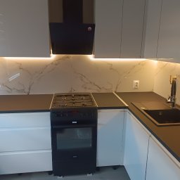 Usługi remontowo budowlane Jakub Justynski - Nowoczesna kuchnia z białymi szafkami, czarnym blatem i płytą gazową Amica, oświetleniem LED pod szafkami wiszącymi i marmurowym wzorem na ścianie.