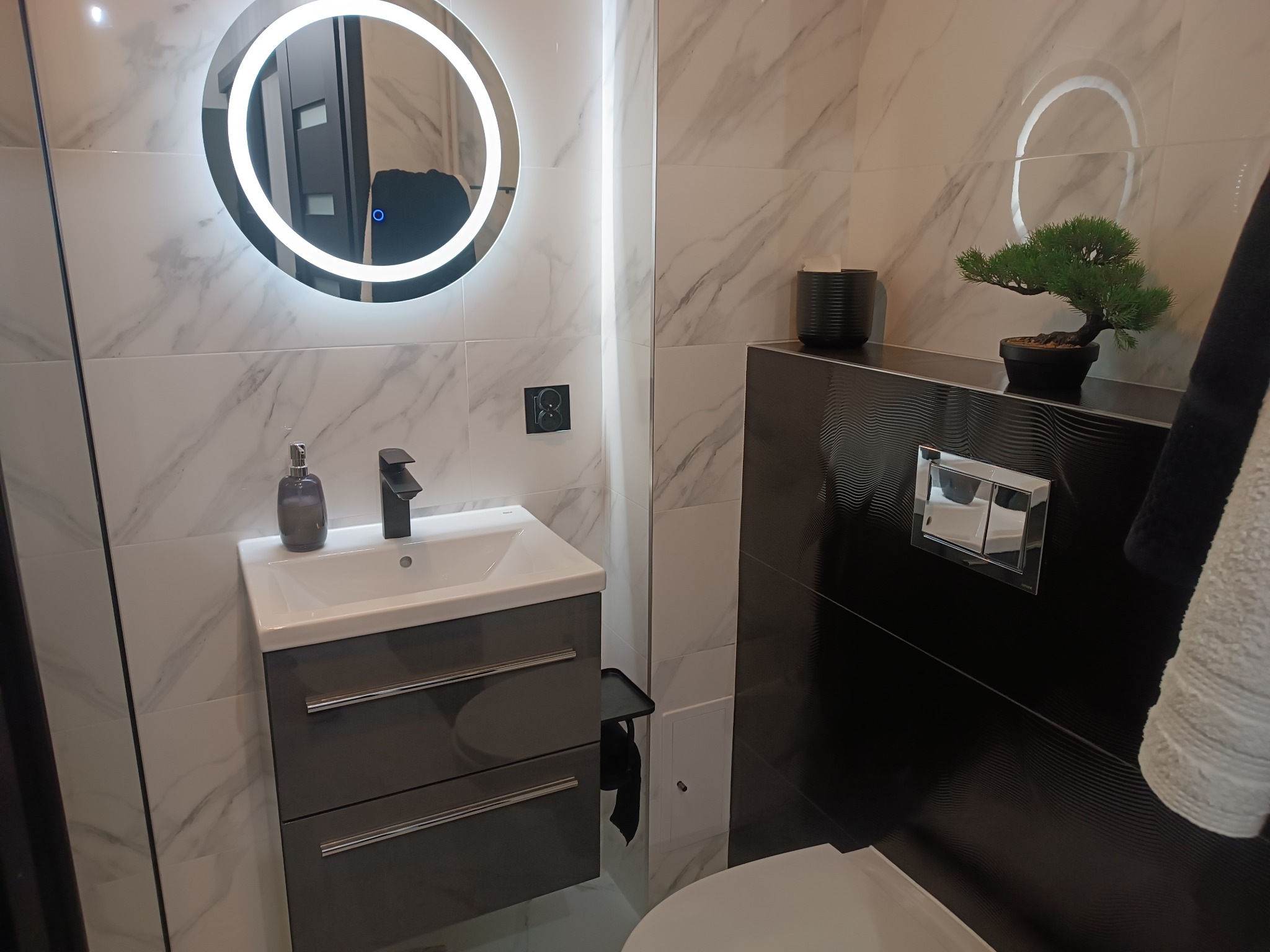 Nowoczesna łazienka z umywalką nablatową na szafce w odcieniach szarości, okrągłe lustro z podświetleniem LED, białe płytki imitujące marmur oraz czarne płytki z falistym wzorem, dekoracyjna...