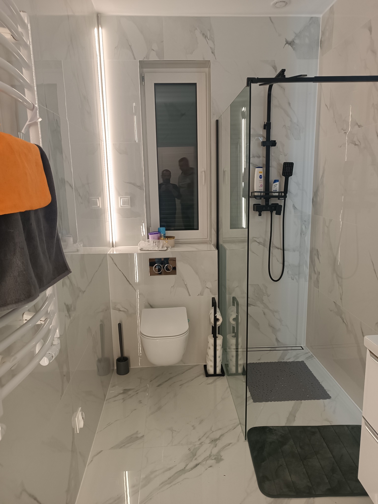 Nowoczesna łazienka wykończona białymi płytkami imitującymi marmur, z kabiną prysznicową typu walk-in z czarnymi elementami, wiszącą toaletą i grzejnikiem drabinkowym z powieszonym pomarańczowym...