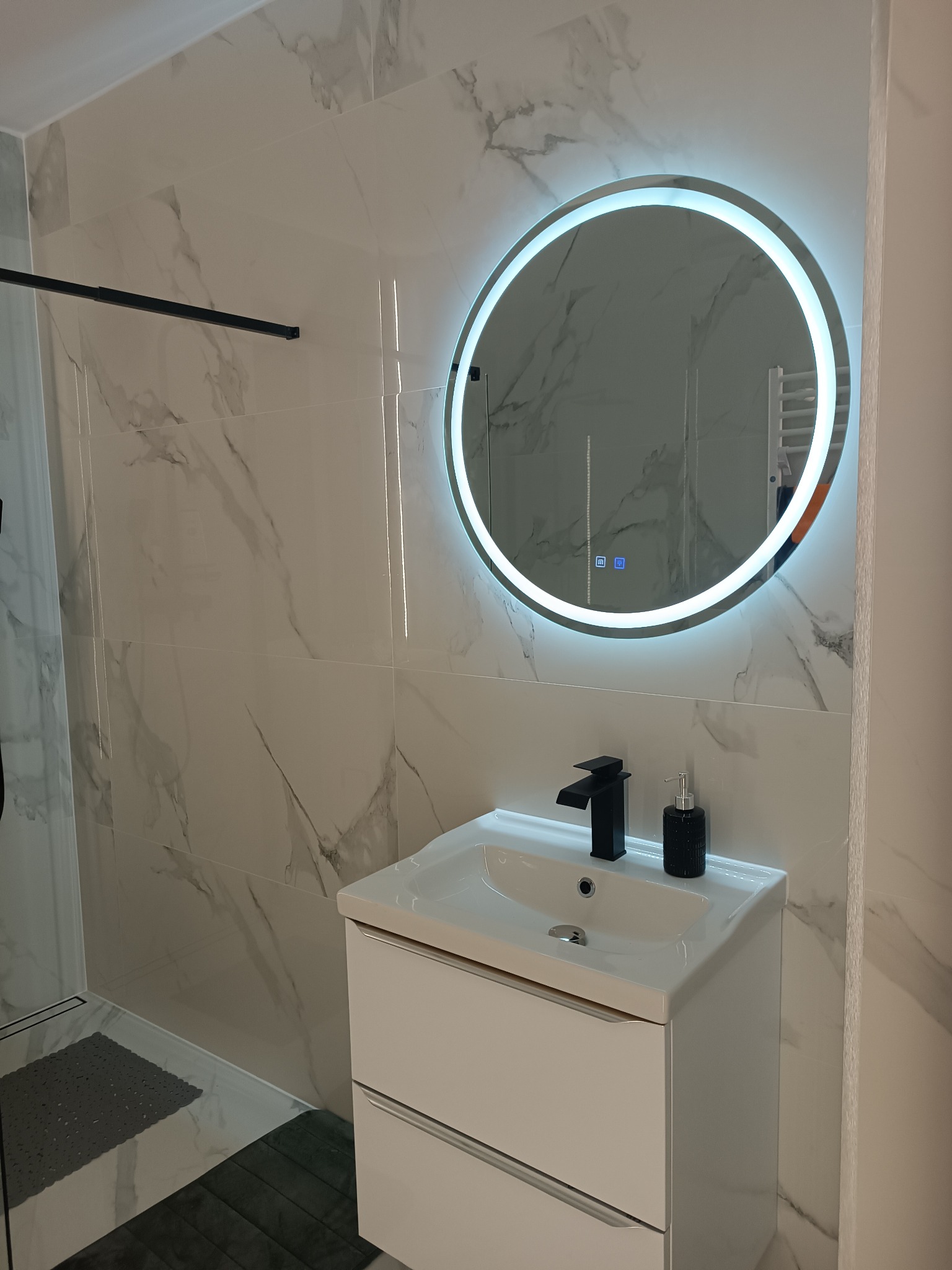 Nowoczesna łazienka z białymi marmurkowymi płytkami, okrągłym lustrem z podświetleniem LED, białą szafką pod umywalką i czarną baterią oraz dozownikiem mydła.