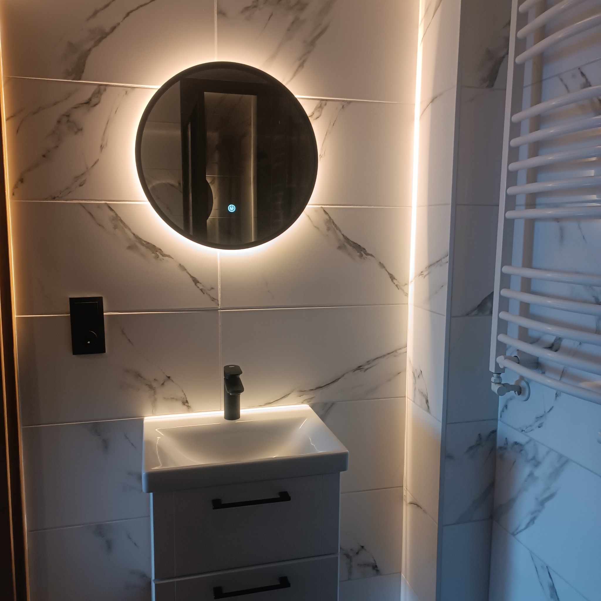 Nowoczesna łazienka z białymi marmurkowymi płytkami, okrągłym podświetlanym lustrem z włącznikiem dotykowym, białą szafką pod umywalkę z czarnymi uchwytami i czarną armaturą oraz białym grzejnikiem...