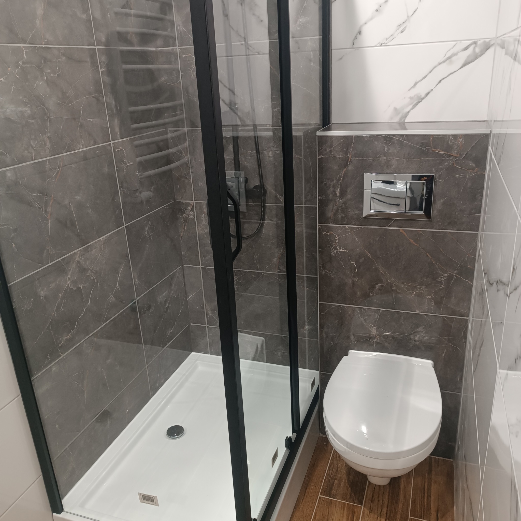 Nowoczesna łazienka z kabiną prysznicową z czarnymi ramami, wiszącą toaletą i płytkami imitującymi marmur w odcieniach szarości i bieli.